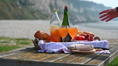 Normandiya 'da Fransız cilası, bölgesel peynir ve taze elmaların yer aldığı samimi bir plaj pikniği manzaralı bir sahilin arka planında geçiyor.