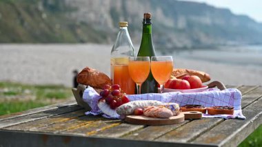 Normandiya 'da Fransız cilası, bölgesel peynir ve taze elmaların yer aldığı samimi bir plaj pikniği manzaralı bir sahilin arka planında geçiyor.