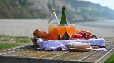 Normandiya 'da Fransız cilası, bölgesel peynir ve taze elmaların yer aldığı samimi bir plaj pikniği manzaralı bir sahilin arka planında geçiyor.