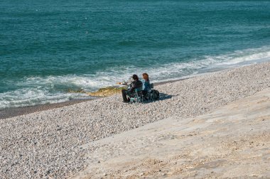 Bir çift, Etretat 'ın çakıllı plajında, manzaralı uçurumlar ve okyanus esintileriyle çevrili, huzurlu bir sahil pikniğinin tadını çıkarıyor..