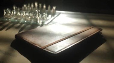 Akşamları masada kahverengi deri bir günlük. Doğal ışık ve arka planda satranç tahtası olan gölgeler.