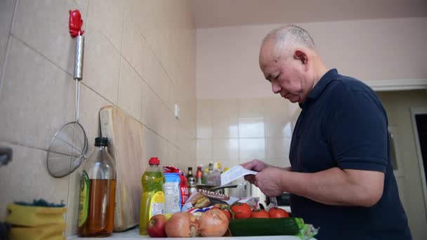Starszy Mężczyzna Kuchni Sprawdza Koszty Codziennej Żywności Artykułów Gospodarstwa Domowego — Wideo stockowe