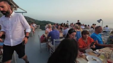 Exanthia, Lefkada, Yunanistan-07.01.2024. Rachi Restoranı 'nda akşam yemeği ve sosyalleşme. Yanında deniz ufkunun altında batan güneşin güzel dramatik dağ manzarası..