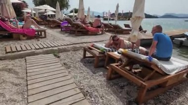 Nidri, Lefkada, Yunanistan-07.02.2024. Kumsalda güneşli yataklarda dinlenen turistler restoran ve barla doğrudan sahilde yiyecek ve içecek sağlıyorlar.. 