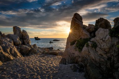 Güneşin denizin üzerinde battığı kayalık bir sahil gökyüzü bulutları üzerine parlayan ışıklar ve bir kayanın arkasından süzülen güneş ışığı. Tatil, ekoloji, çevre ve doğa geçmişi.
