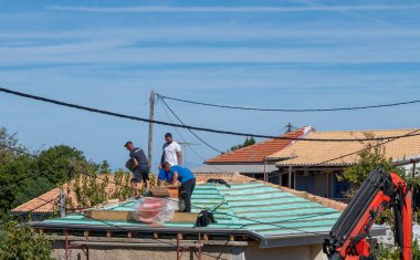 Lefkada, Yunanistan... 10.08.2024. İnşaat işçileri tadilat altındaki bir binanın tepesinde çatıyı seramik fayanslarla kaplıyorlar..