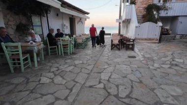 Lefkada. Yunanistan... 10.16.2024. Akşamları Agios Nikitas sahiline doğru yürüyen bir çift diğer turistlerle fresk yemeğinin tadını çıkarıyor..