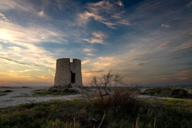 Gün batımında, güneş ışığı ve gökyüzü bulutlarıyla kumsalda eski bir yel değirmeni..