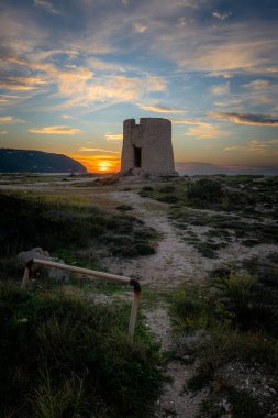 Gün batımında, güneş ışığı ve gökyüzü bulutlarıyla kumsalda eski bir yel değirmeni..