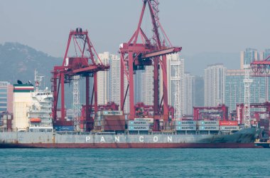 Hong Kong. Çin... 02.18.2025. Hong Kong Limanı 'nın arka planında rıhtımdaki vinçler ve yüksek binalarla kenetlenmiş bir kargo gemisinin görüntüsü..