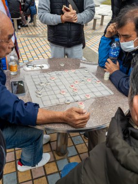 Hong Kong. Çin - 02.23.2025. Sha Tin Park 'ta Çin satrancı oynayan iki yaşlı adamın etrafı izleyicilerle çevrili..
