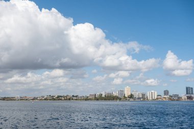 Güney Perth, Avustralya manzaralı. Ön planda durgun mavi bir nehir ve mavi gökyüzü altında tüylü beyaz bulutları olan modern yerleşim ve ticari binaların ufuk çizgisini gösteriyor.