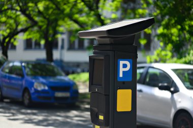 Parlak bir günde park halindeki arabaların bitişiğindeki yeşil ağaçlarla dolu bir caddede güneş enerjisiyle çalışan parkmetre belirgin bir şekilde duruyor..