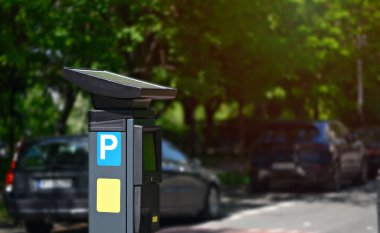 Güneşli bir günde, ağaçlık bir sokak boyunca park halindeki arabalarla çevrili modern bir parkmetre görülebilir..