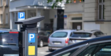 Güneşli bir günde şehrin yakınına park edilmiş birden fazla aracın park ettiği bir parkmetre belirgin bir şekilde sergilenir..