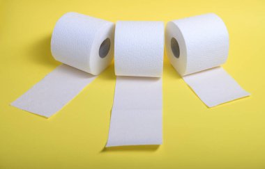 Sarı bir yüzeyde sergilenen üç rulo yumuşak beyaz tuvalet kağıdı, her biri aşağı sarkıyor..