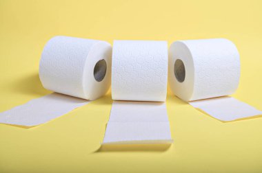 Üç rulo yumuşak beyaz tuvalet kağıdı parlak sarı arka planda oturuyor, hijyeni vurguluyor..