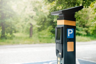 Yol kenarında ağaçlarla çevrili bir parkmetre var. Güneşli bir günde huzurlu bir şehir ortamı olduğunu gösteriyor..