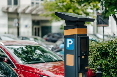 Parlak güneş ışığı altında park halindeki araçlarla dolu kentsel bir ortamda güneş enerjili bir parkmetre bulunmaktadır..