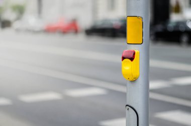 Sarı bir trafik sinyal kontrol düğmesi, gri bir direğe yerleştirilmiş, şehirde arabalarla dolu bir yolun kenarında, gün ışığında yakalanmış..