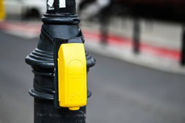 Parlak sarı bir yaya sinyal düğmesi, işlek bir kentsel alanda yaya geçidini gösteren bir elektrik direğine iliştirilmiş, kullanıma hazır..