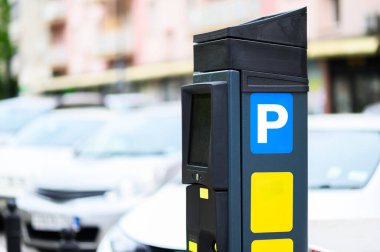 Modern bir parkmetre, kalabalık bir caddede çeşitli park edilmiş araçlarla çevrili kentsel bir çevrede duruyor..