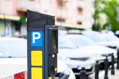 Canlı bir şehir bölgesinde park halindeki birkaç aracın yanına günlük trafiği ve şehir hayatını gösteren bir parkmetre yerleştirildi..