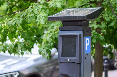 Güneş enerjisiyle çalışan bir parkmetre yemyeşil yeşilliklerin altına yerleştirilmiş, kentsel bir ortamda çevre dostu teknolojiyi vurguluyor..
