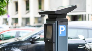 Güneş enerjisiyle çalışan parkmetre, park halindeki araçlarla dolu, modern altyapıyı gözler önüne seren işlek bir kentsel alanda duruyor..