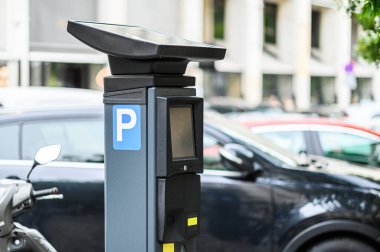 Güneş panelleriyle donatılmış bir parkmetre, yoğun bir şehir bölgesinde gün ışığında park halindeki arabaların arasında duruyor..