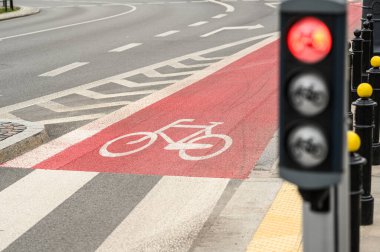 Kalabalık bir şehir kavşağında, kentte bisiklet güvenliğini vurgulayan kırmızı bir bisiklet yolu ve trafik ışıkları görülüyor..