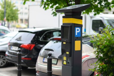 Güneş panelleriyle donatılmış modern bir parkmetre çeşitli park araçları ve yeşilliklerle çevrili kentsel bir ortamda duruyor..