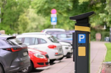 Kalabalık bir kentsel alanda park halindeki birkaç arabanın yanında duran bir parkmetre arka planda açık tabela ve yeşillik gösteriyor..