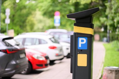 Yeşil ağaçlar ve güneş ışığıyla çevrili bir şehir otoparkında birkaç aracın yanına bir parkmetre yerleştirildi..
