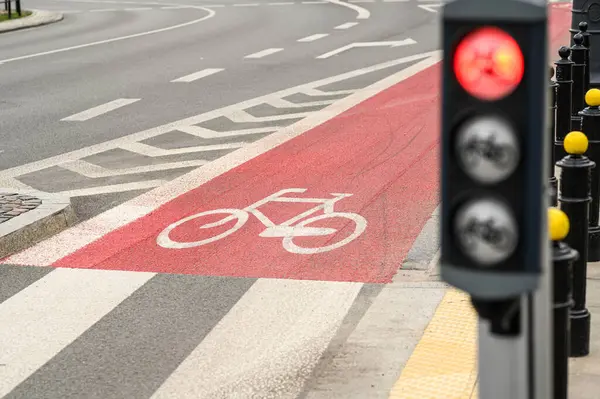 Kalabalık bir şehir kavşağında, kentte bisiklet güvenliğini vurgulayan kırmızı bir bisiklet yolu ve trafik ışıkları görülüyor..