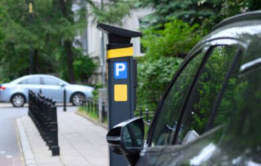 Şehir caddesinde birçok park etmiş aracın yanında gün ışığında yeşilliklerle çevrili bir park makinesi bulunuyor..