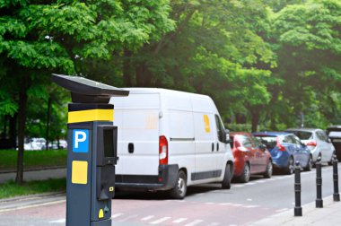 Ağaçlık bir caddede, çeşitli arabaların yanında bir parkmetre duruyor. Şehir ulaşımını ve güneşli havayı gözler önüne seriyor..