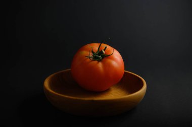 Canlı kırmızı renkte olgun bir domates, pürüzsüz bir ahşap kasede oturur. Koyu bir arka planda aydınlanır ve tazeliğini gösterir..