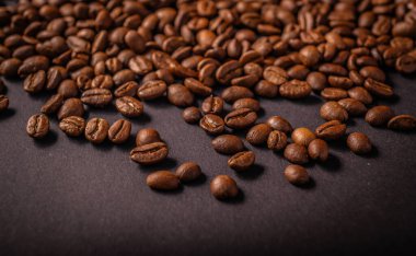 Taze kavrulmuş kahve çekirdekleri koyu bir arkaplana saçılmış, parlak dokuları ve koyu kahverengi renkleri vurgulanıyor..