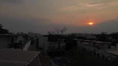 Bir kadın alacakaranlıkta beyaz evlerin üzerine düşerken dramatik günbatımında çatı bahçesinde vakit geçiriyor. Kamerayı yukarı eğ.