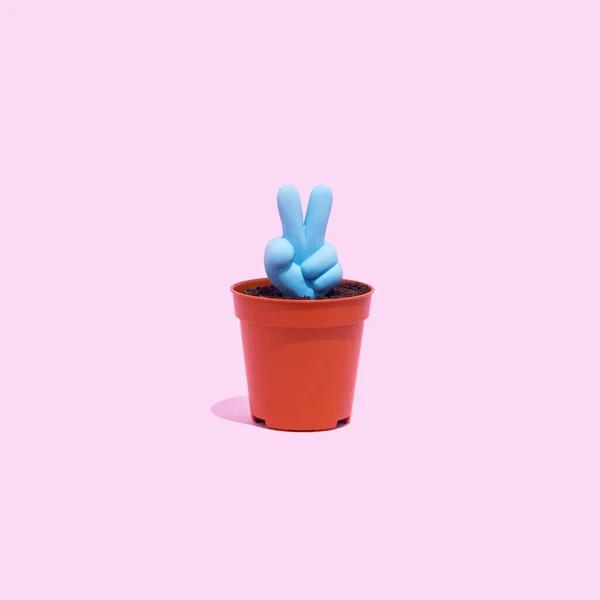 Blue peace, victory symbol icon growing from a small pot on a pastel pink background. Hand gesture, peace or victory concept.
