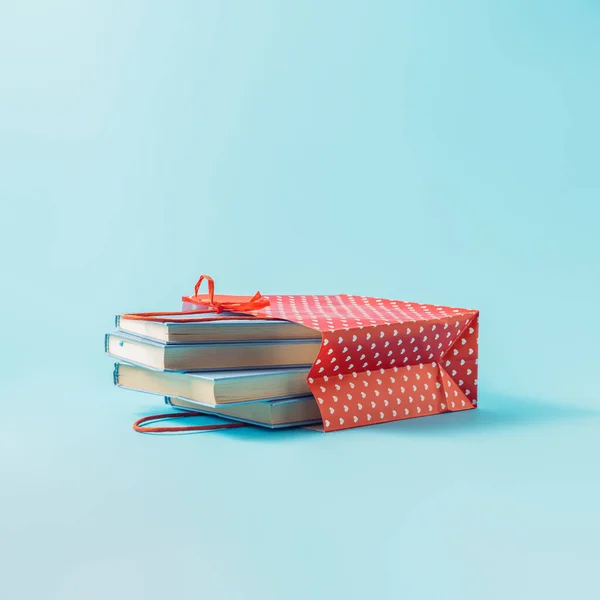 Aesthetic composition of pastel blue books in a red shopping bag and white heart-shaped dots. Concept of buying and selling books, gift books, education.