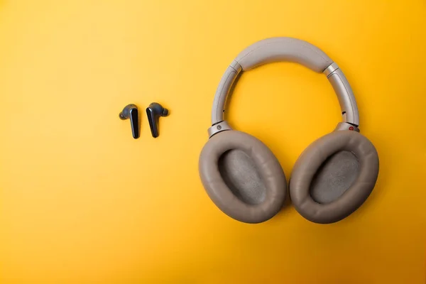 Light gray wireless on-ear headphones and blue in-ear wireless headphones on a yellow background. Comparison of different types of headphones. Headphones for playing games or listening to music. Noise canceling headphones. Top view. Copy space.