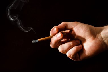 Bir adamın eli koyu bir arka planda yanan bir sigarayı tutar. Sert ışık. Sigara içiyorum. İnsanların kötü alışkanlıkları. Akciğer kanseri. Sigaraya karşı savaş