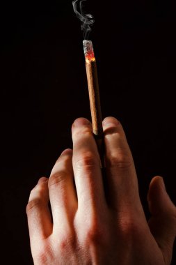 Bir adamın eli koyu bir arka planda yanan bir sigarayı tutar. Sert ışık. Sigara içiyorum. İnsanların kötü alışkanlıkları. Akciğer kanseri. Sigaraya karşı savaş