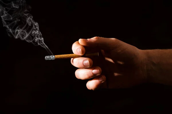 A man\'s hand holds a lit cigarette on a dark background. Hard light. Smoking. Bad habits of people. Lungs\' cancer. Fight against smoking
