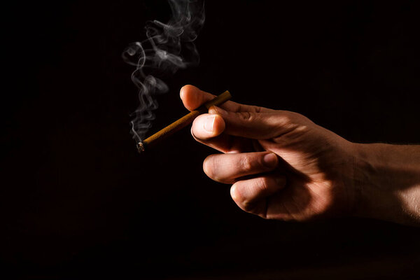 Мужская рука держит зажженную сигарету на темном фоне. Жесткий свет. Курение. Плохие привычки людей. Рак легких. Борьба с курением