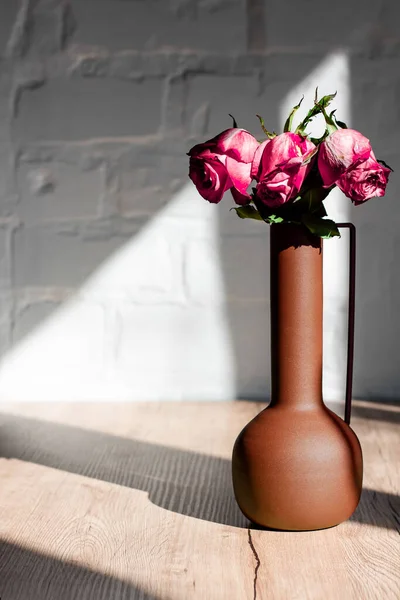 Withered pink roses in a vase in the hard rays of the sun on a table against a background of white brick.