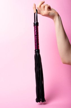 Kadın eli pembe arka planda pembe saplı siyah deri bir kırbaç tutuyor. Mesaj için yer. Seks dükkanı konsepti. Boşluğu kopyala.