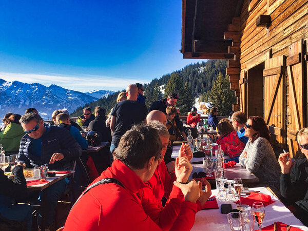 manigod, france - january 13 2018 : many people are siting in the sun eating and drinking on the terrace of a wood log restaurant